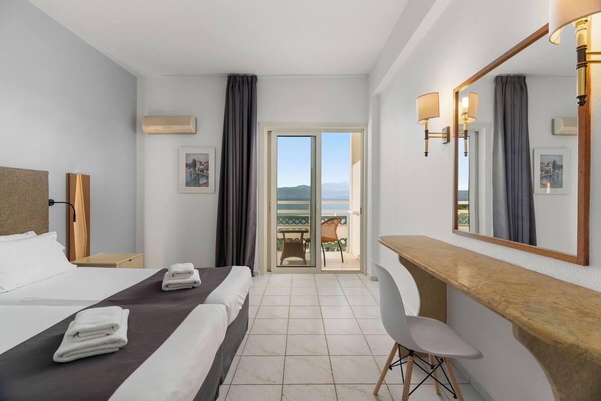 A hotel room with two beds, a desk, a chair, a large mirror, and a view of the sea through a sliding door.