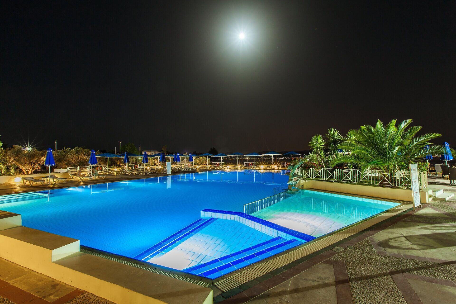 A nighttime swimming pool with blue lighting, lounge chairs, and umbrellas.