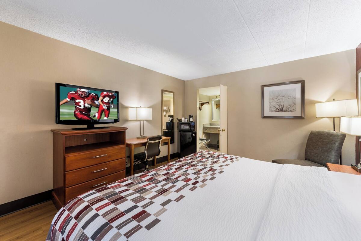 A hotel room with a large bed, a desk with a chair, a television, a refrigerator, and a mirror.