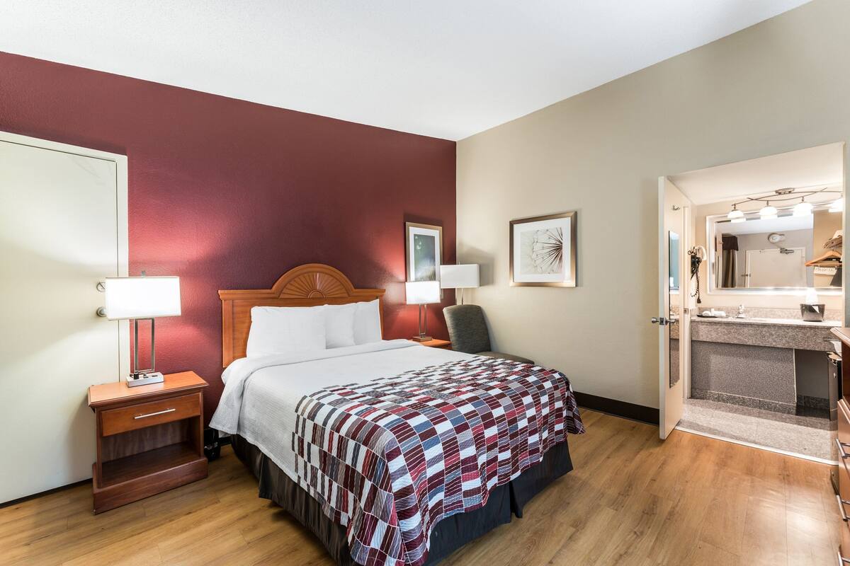 A hotel room with a bed, bedside tables, a nightstand, a desk, a chair, a desk lamp, a mirror, and a bathroom with a sink and mirror.