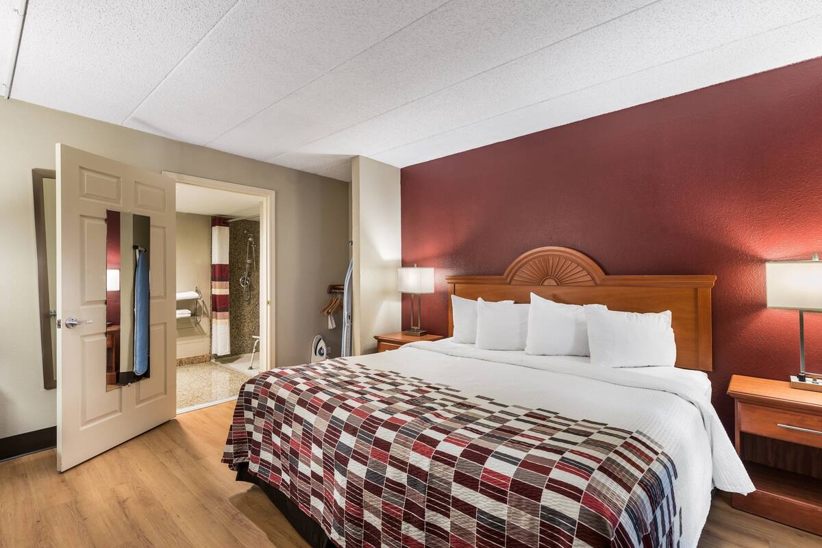 A hotel room with a large bed, a wooden headboard, a red accent wall, a wooden floor, and a bathroom visible through an open door.