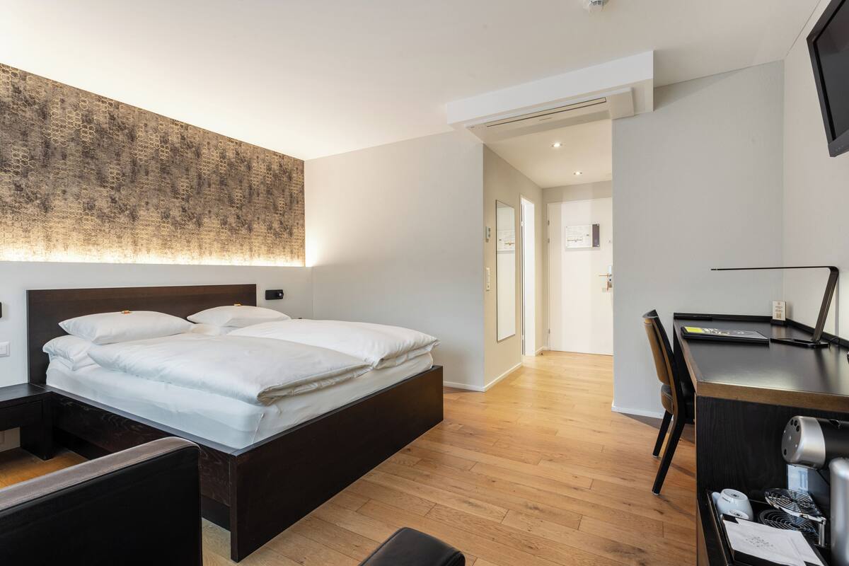 Quarto de hotel moderno com uma cama grande, uma escrivaninha e uma cadeira. O chão é de madeira e há uma grande decoração na parede.