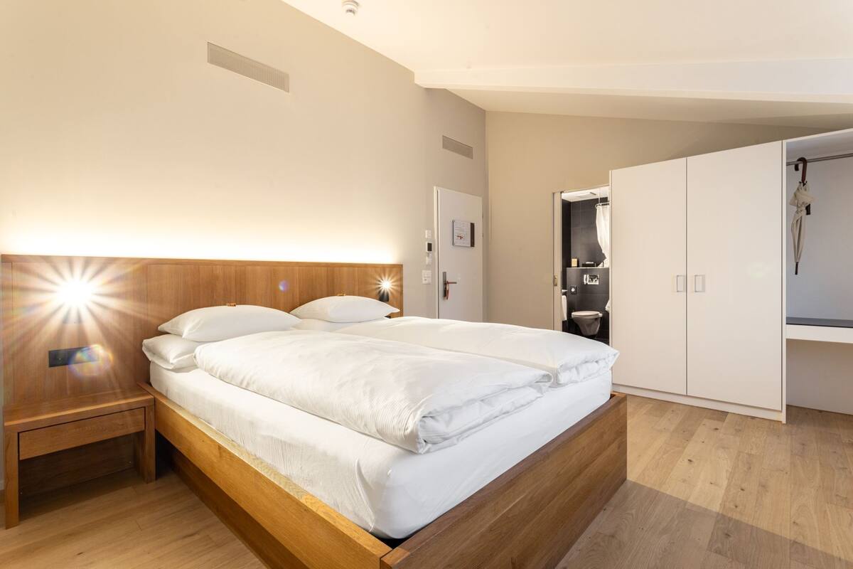 Quarto moderno com cama de madeira, mesa de cabeceira, guarda-roupa e banheiro visível através de uma porta aberta.
