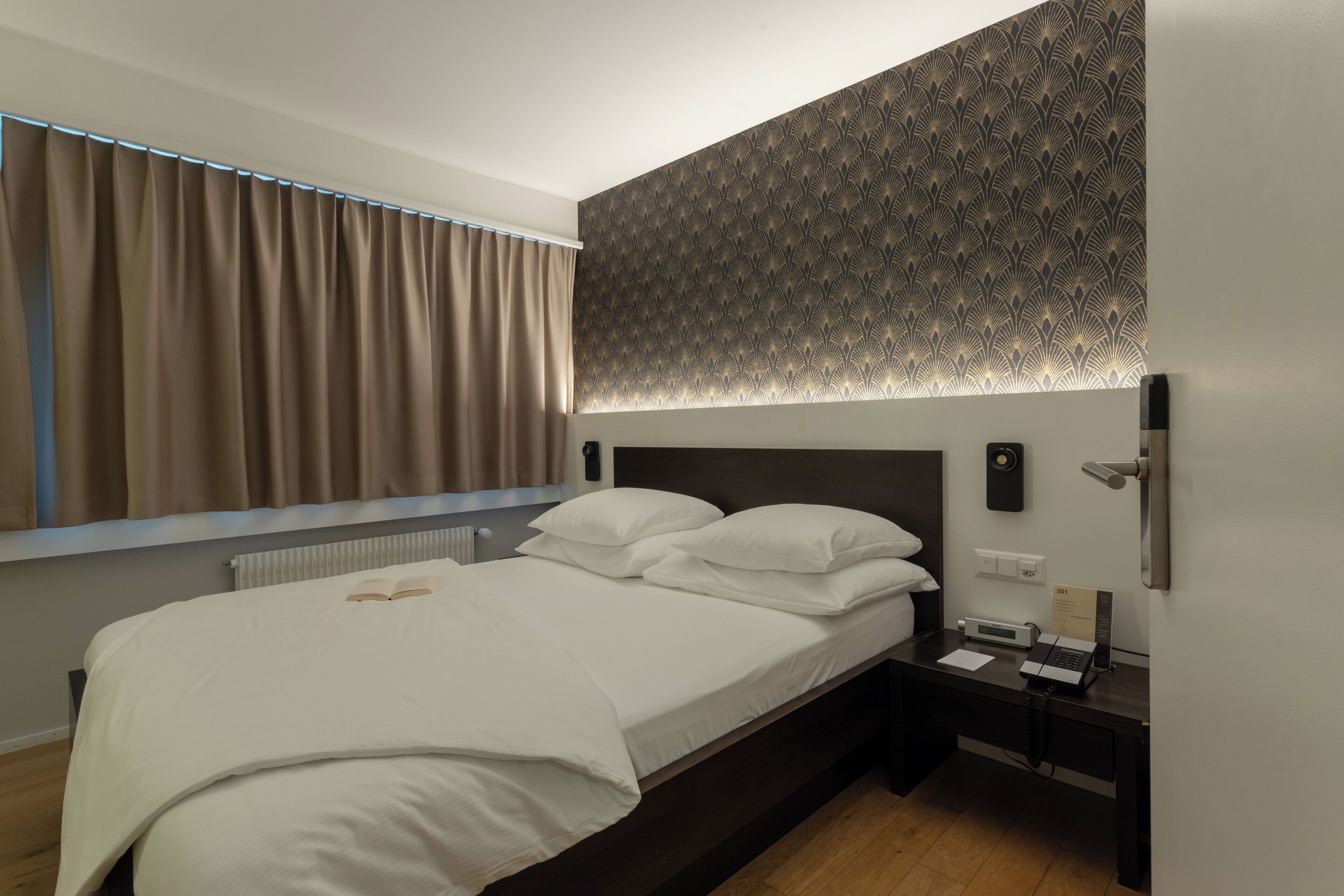 A neatly made bed with white linens, a bedside table with a phone and a small lamp, and a patterned wall above the bed.