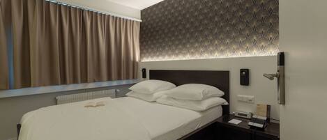 A neatly made bed with white linens, a bedside table with a phone and a small lamp, and a patterned wall above the bed.