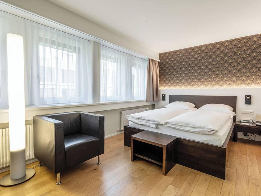 Una habitación de hotel moderna con un ventanal grande, una cama, un sofá, una mesita de noche y un aplique de pared.