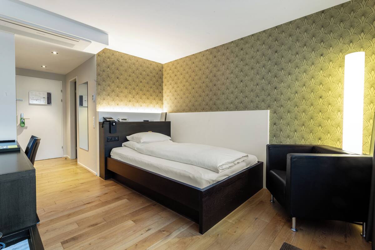 Quarto moderno com piso de madeira, cama com roupa de cama branca, cabeceira preta, poltrona preta e papel de parede estampado.