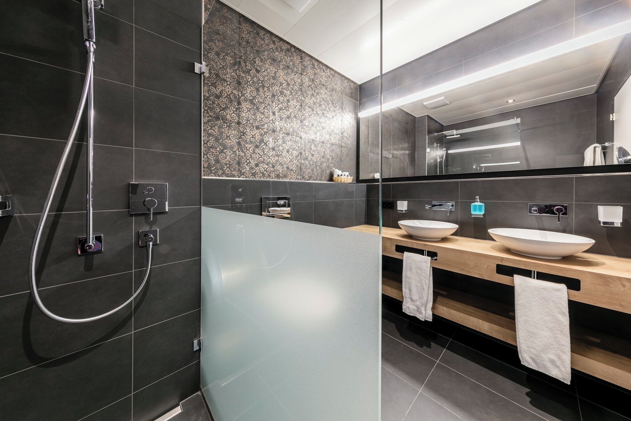 Une salle de bain moderne avec des carreaux de couleur sombre, une douche en verre et deux vasques avec des cuves blanches.