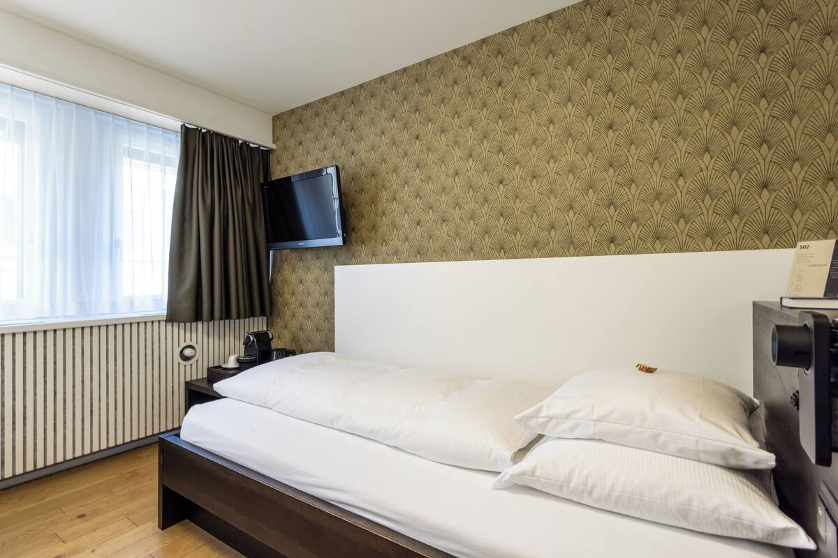 Quarto de hotel com cama, televisão presa à parede, janela com cortinas e radiador.