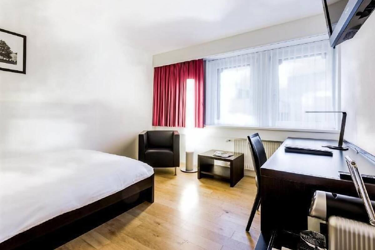 Quarto de hotel com uma cama, uma escrivaninha, uma cadeira, uma mesinha e um radiador.