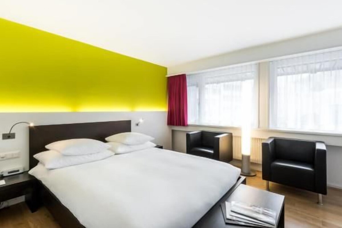 Une chambre d’hôtel avec un lit, des tables de chevet, un bureau, une chaise et une fenêtre avec des rideaux.