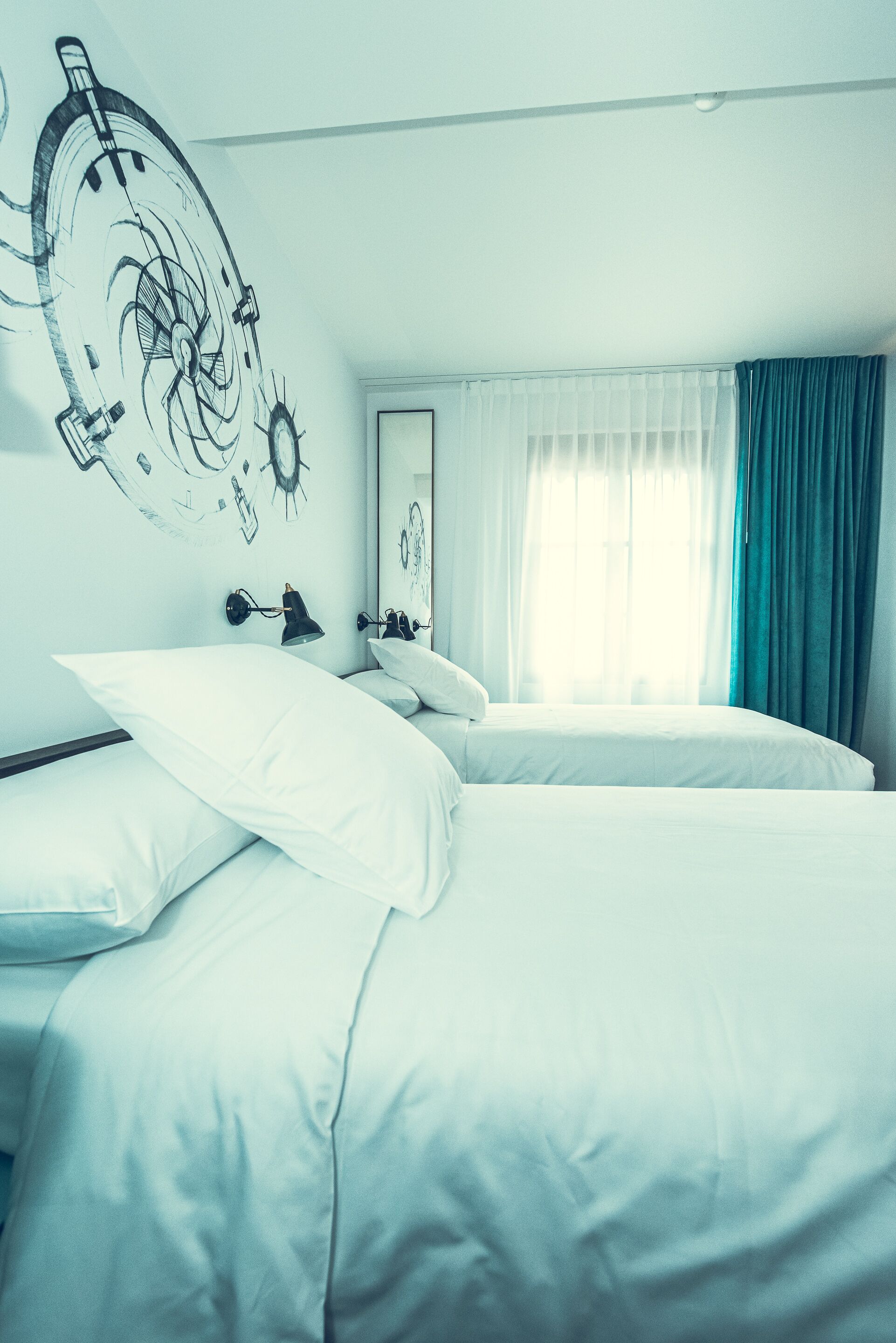A hotel room with two beds, a large wall clock, a window with curtains, and a mirror.
