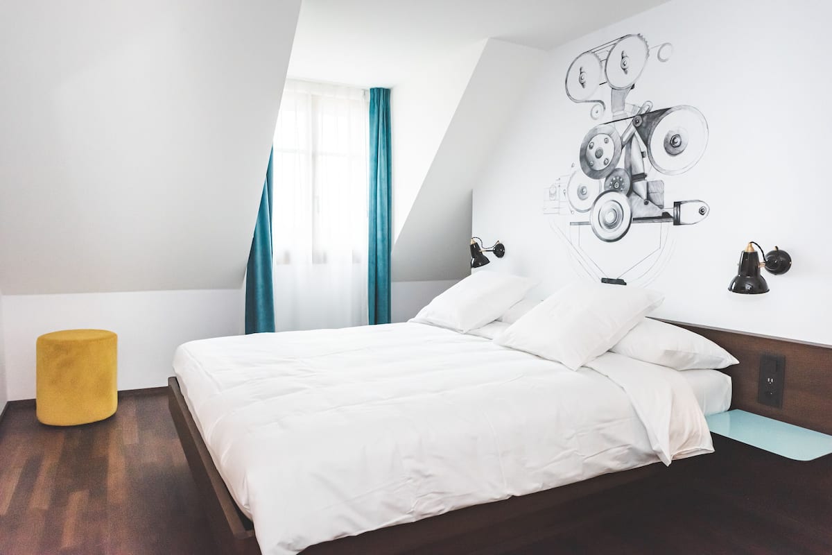A modern hotel room with a bed, a yellow ottoman, a wall-mounted lamp, and a large abstract painting.