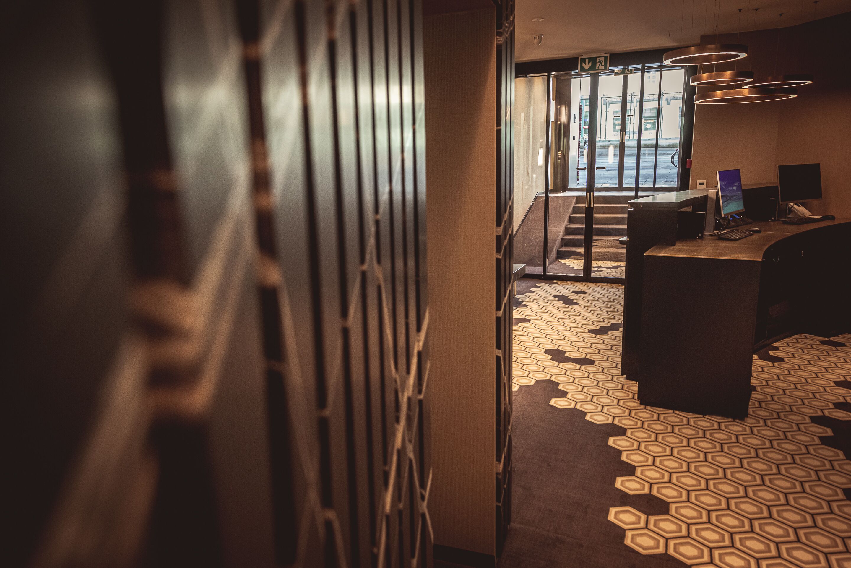 Un vestíbulo de hotel con alfombra de diseño geométrico, un mostrador de recepción y una escalera que conduce a un área exterior.