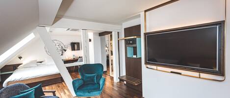 A modern loft bedroom with a large bed, a teal armchair, a TV, and a wooden floor.