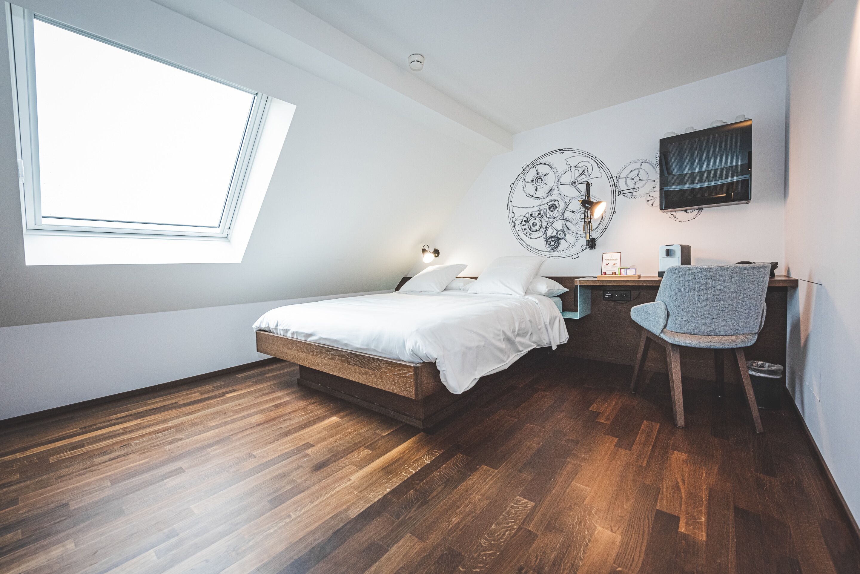 A bedroom with a skylight, a bed, a desk with a chair, and a wall-mounted TV.