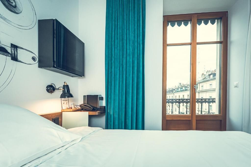 A hotel room with a bed, a desk with a lamp, a telephone, a TV mounted on the wall, and a window with a view of buildings.