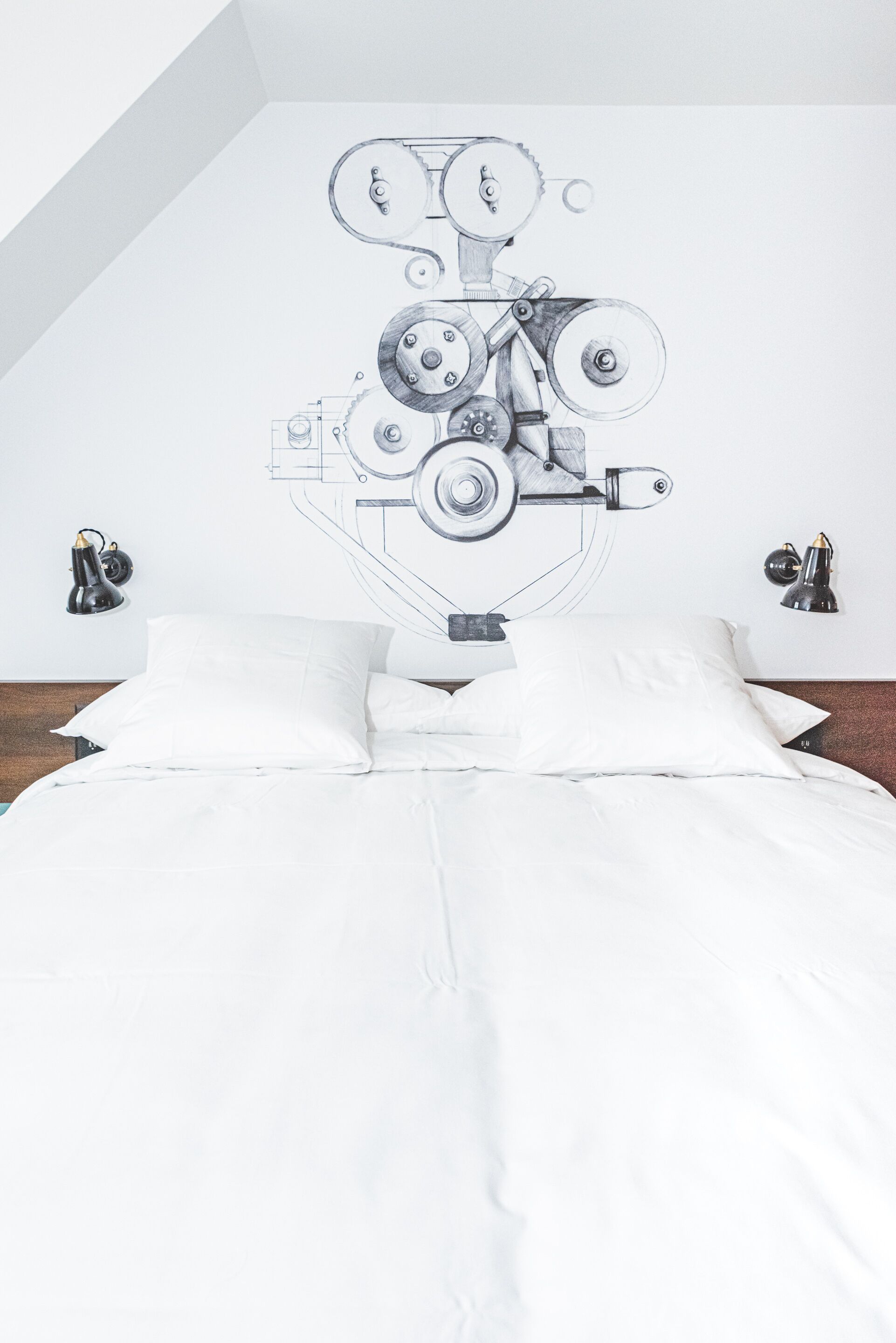 A bedroom with a bed, white bedding, a wall-mounted mechanical drawing, and two wall-mounted lamps.