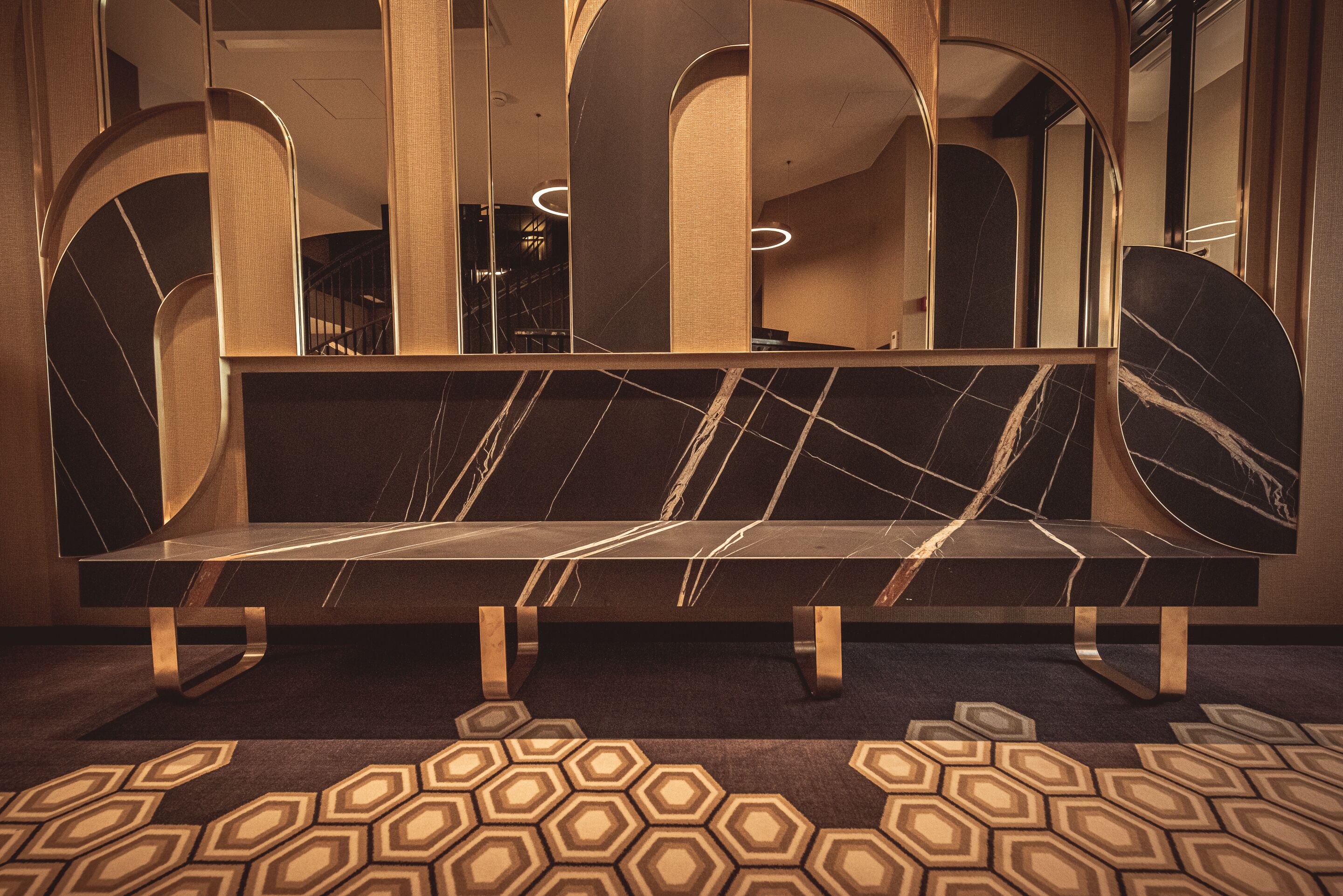 A modern hotel lobby with a marble bench, geometric patterned carpet, and arched mirrors.