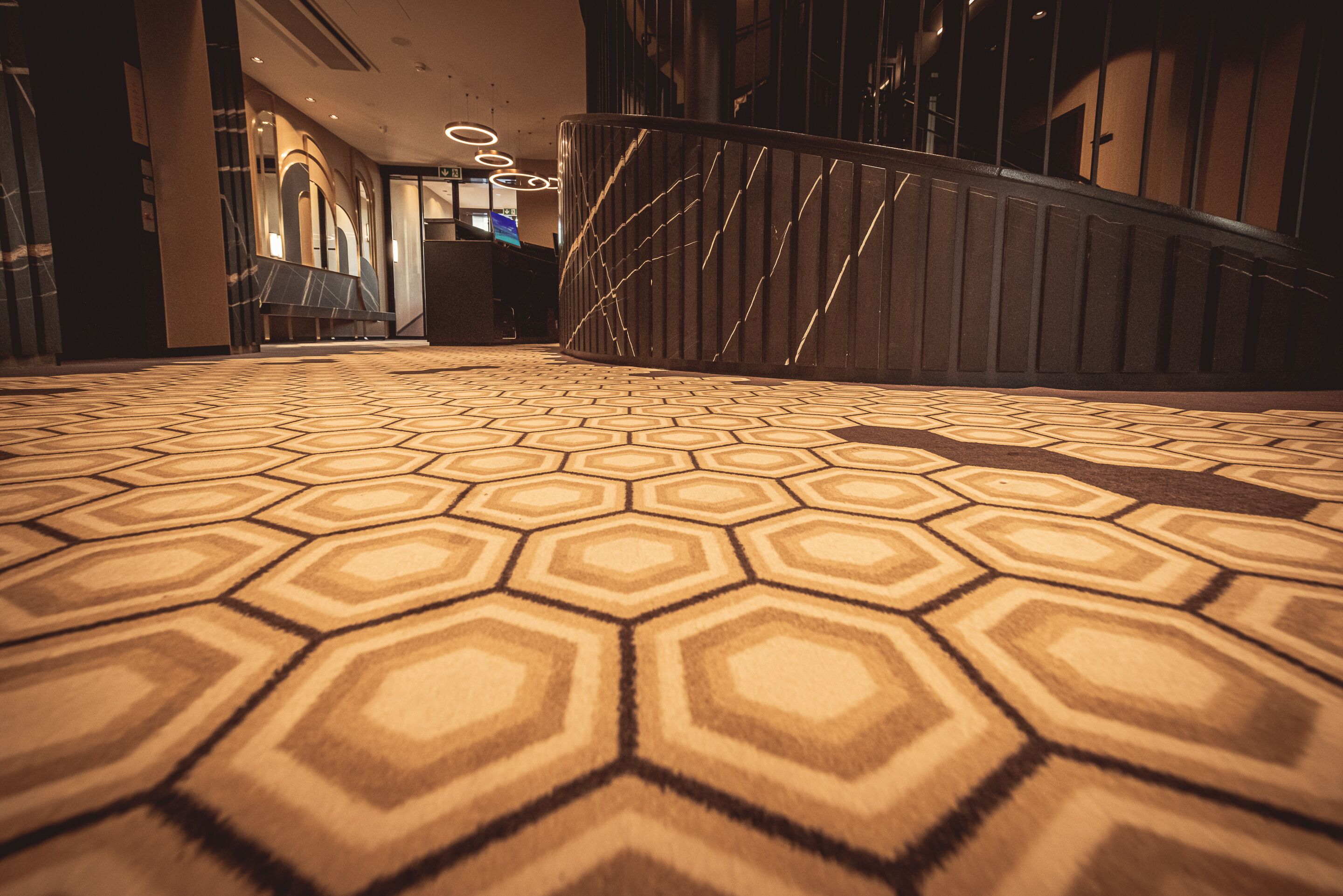 Un hall d’hôtel avec un tapis à motifs géométriques, une rambarde noire incurvée et un comptoir de réception en arrière-plan.