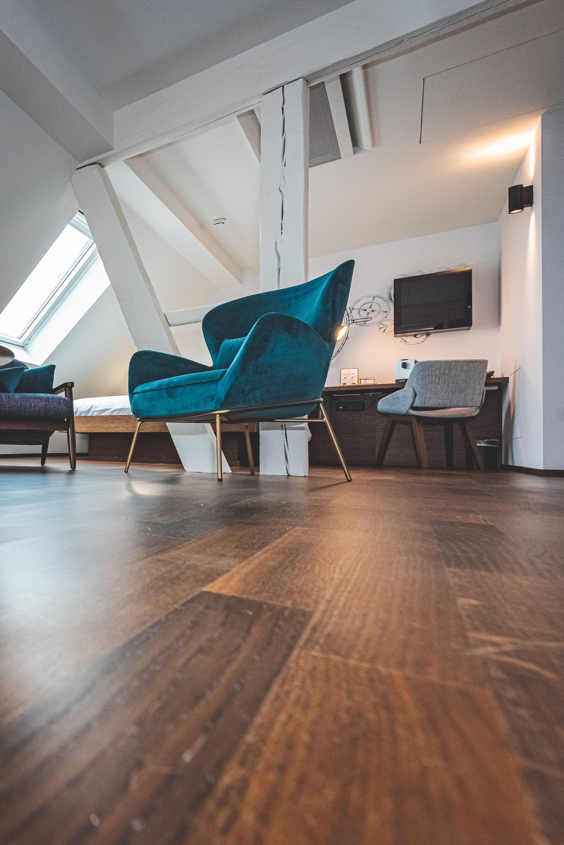 Une chambre de style loft moderne, avec un plafond en pente, une lucarne, un lit, un bureau et un fauteuil de couleur turquoise.