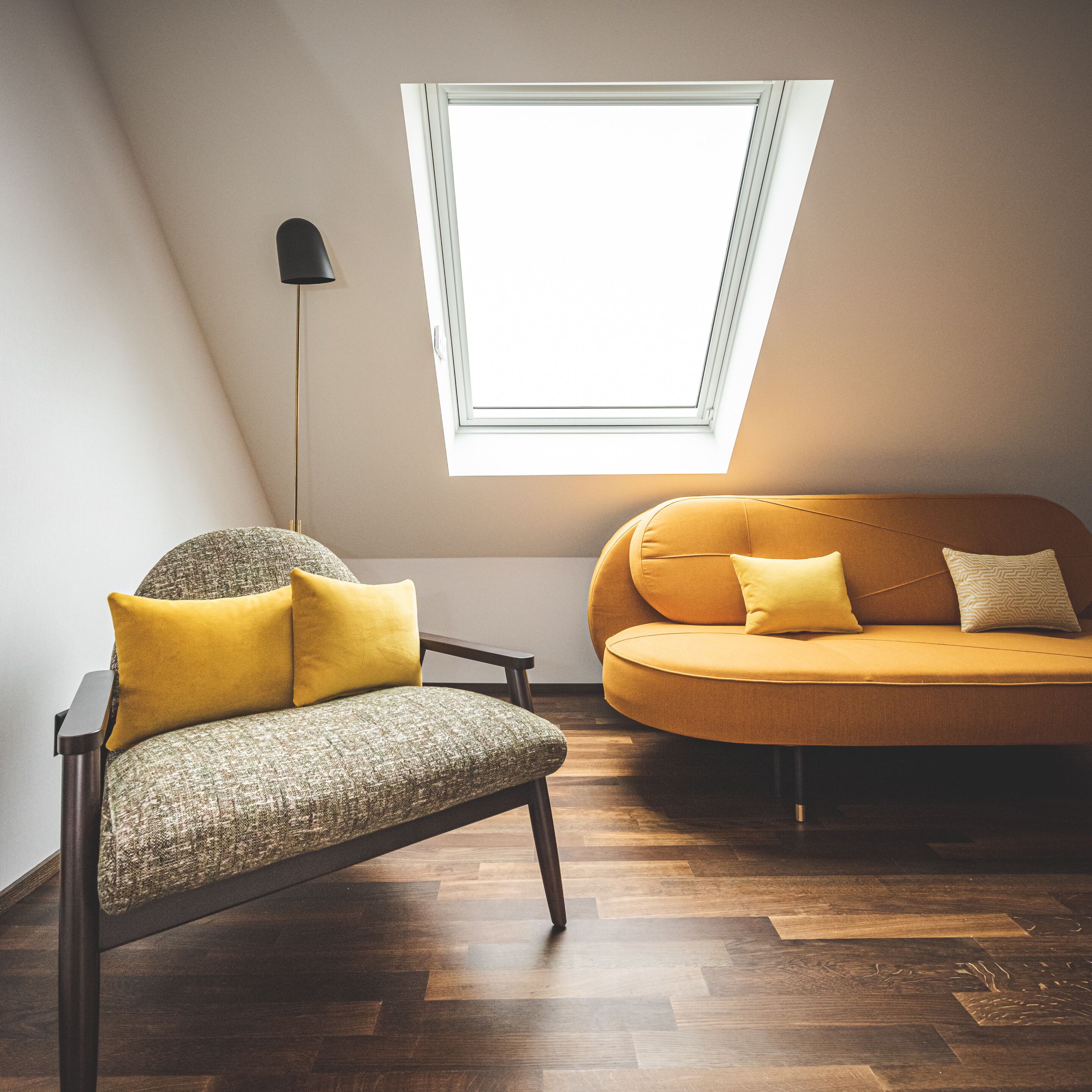 Une chambre de style loft moderne, avec un plafond en pente, un puits de lumière, un fauteuil vert agrémenté de coussins jaunes et un canapé de couleur moutarde avec des coussins à motifs.