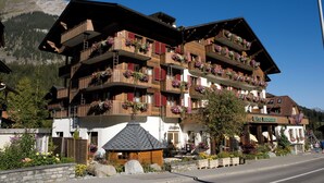 Un hotel de varios pisos con balcones decorados con flores, una señal que indica que es un hotel, y un paisaje montañoso de fondo.