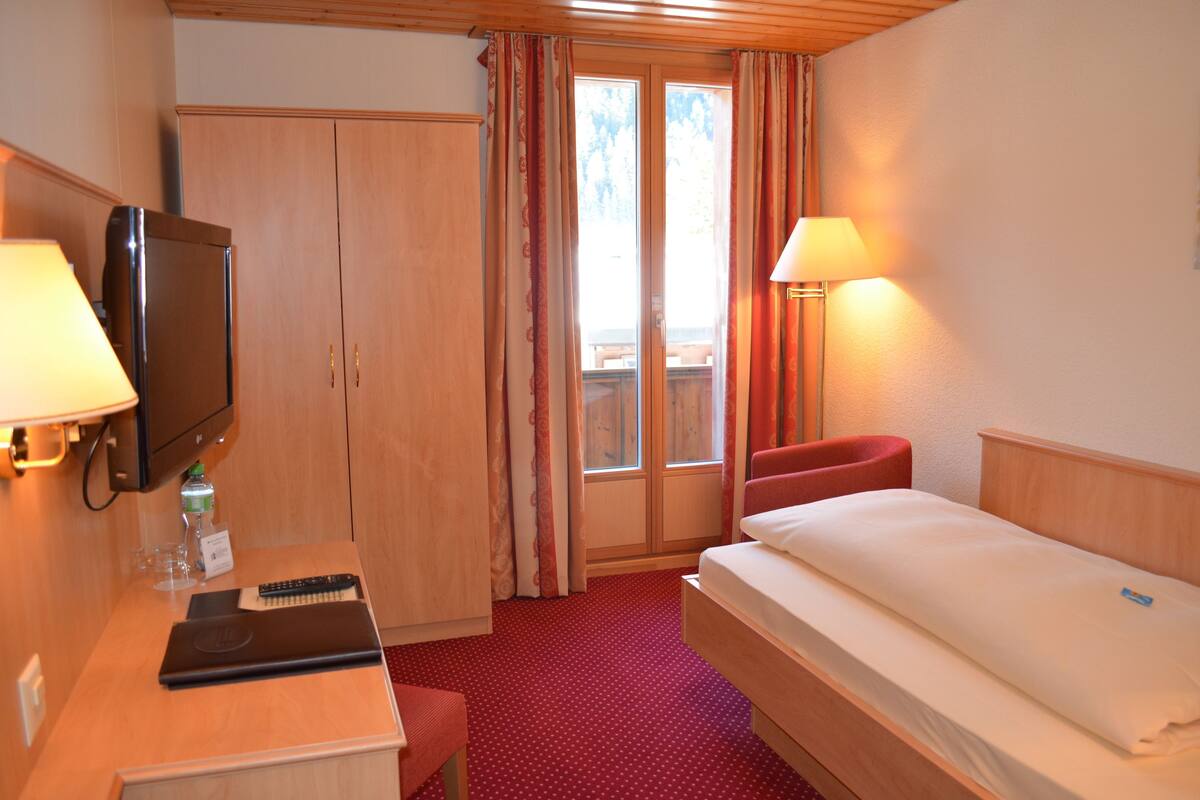 A hotel room with a bed, a desk with a laptop, a TV, and a window with curtains.