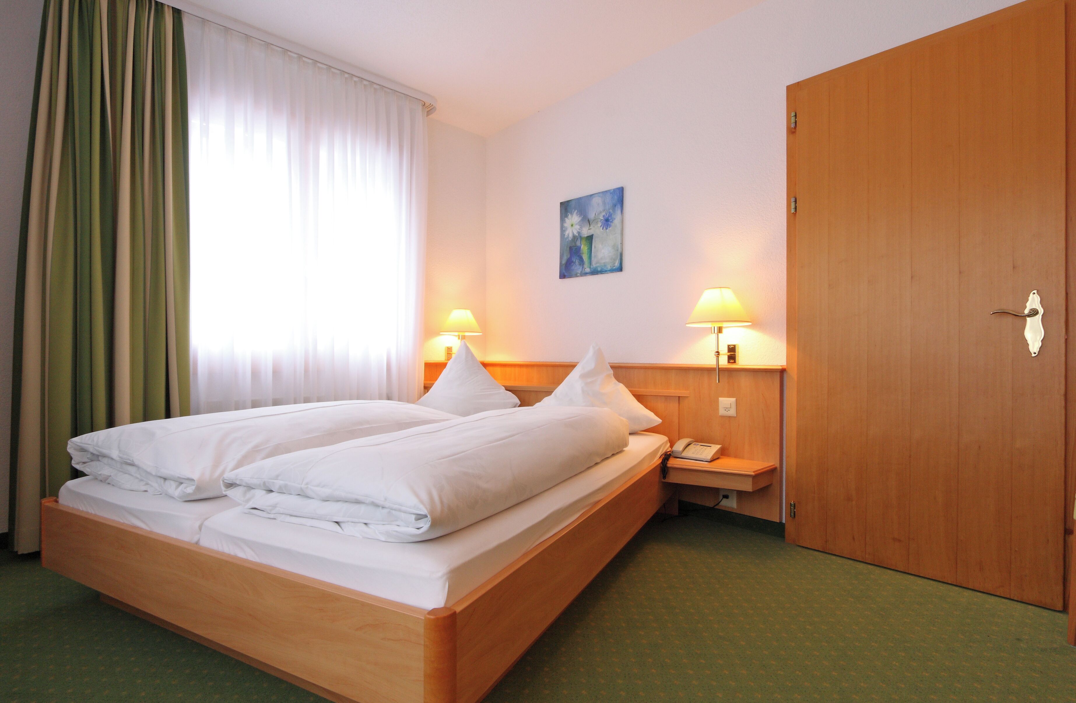 Habitación de hotel con cama doble, dos lámparas de noche, un armario de madera, un cuadro en la pared y una ventana con cortinas.