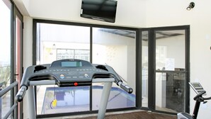 Una caminadora en una habitación con ventanas amplias, un televisor en la pared y vista a un edificio desde el exterior.