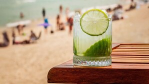 A glass of drink with lime on a wooden table with a beach and mountains in the background. - Hotel Arpoador (Rio de Janeiro)