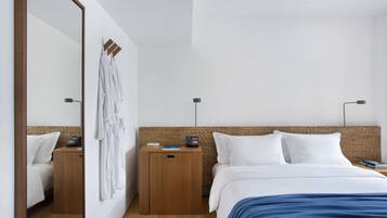 Una habitación de hotel moderna con un espejo grande, una cama con ropa de cama blanca, una manta azul, una mesita de noche de madera y un cabecero de mimbre.