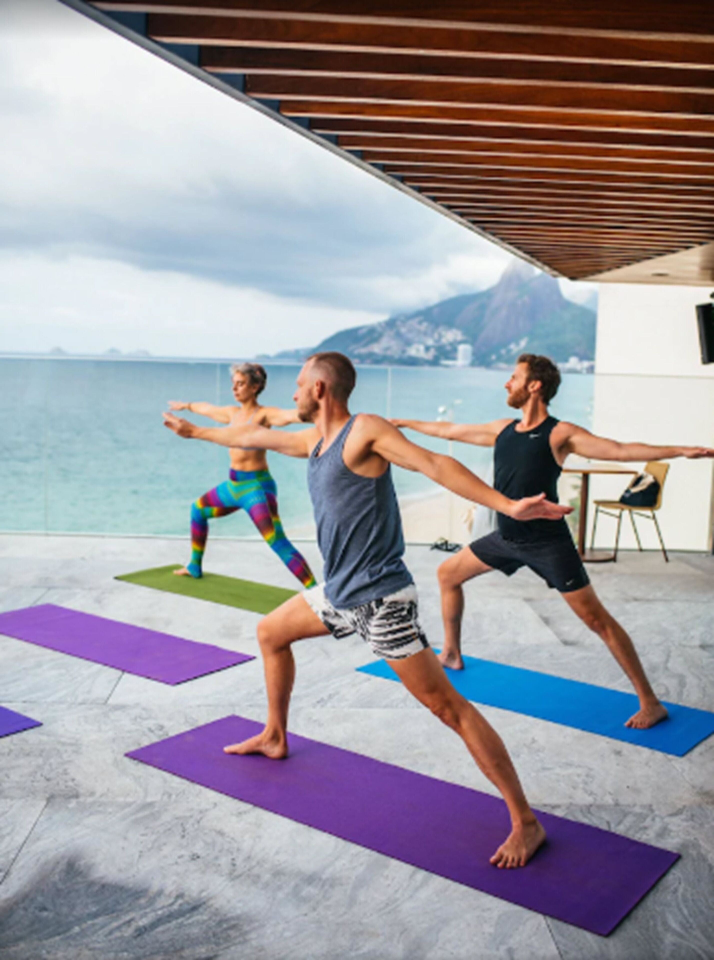 Trois personnes pratiquent le yoga sur des tapis dans un espace extérieur, avec vue sur la mer et les montagnes.