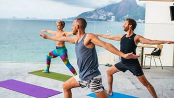 Trois personnes pratiquent le yoga sur des tapis dans un espace extérieur, avec vue sur la mer et les montagnes.
