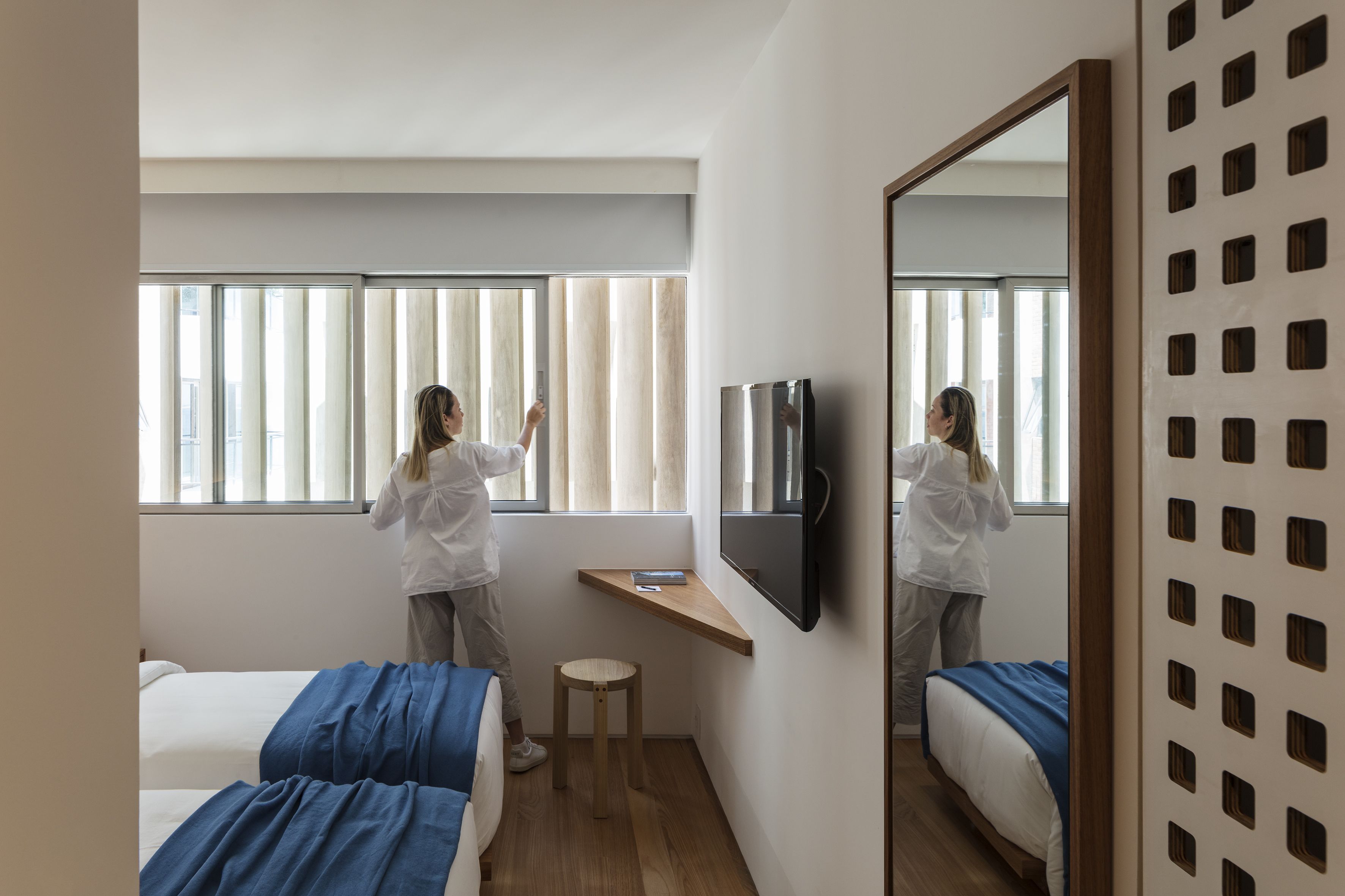 Une chambre d’hôtel avec deux lits, un petit tabouret en bois, un téléviseur fixé au mur et une fenêtre avec des stores.