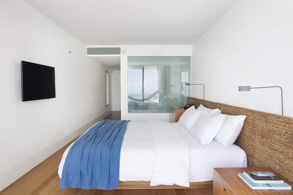 Ein modernes Hotelzimmer mit einem großen Bett, einem Flachbildfernseher an der Wand, einem Nachttisch mit einem Buch und Blick ins Freie durch eine Glastür.