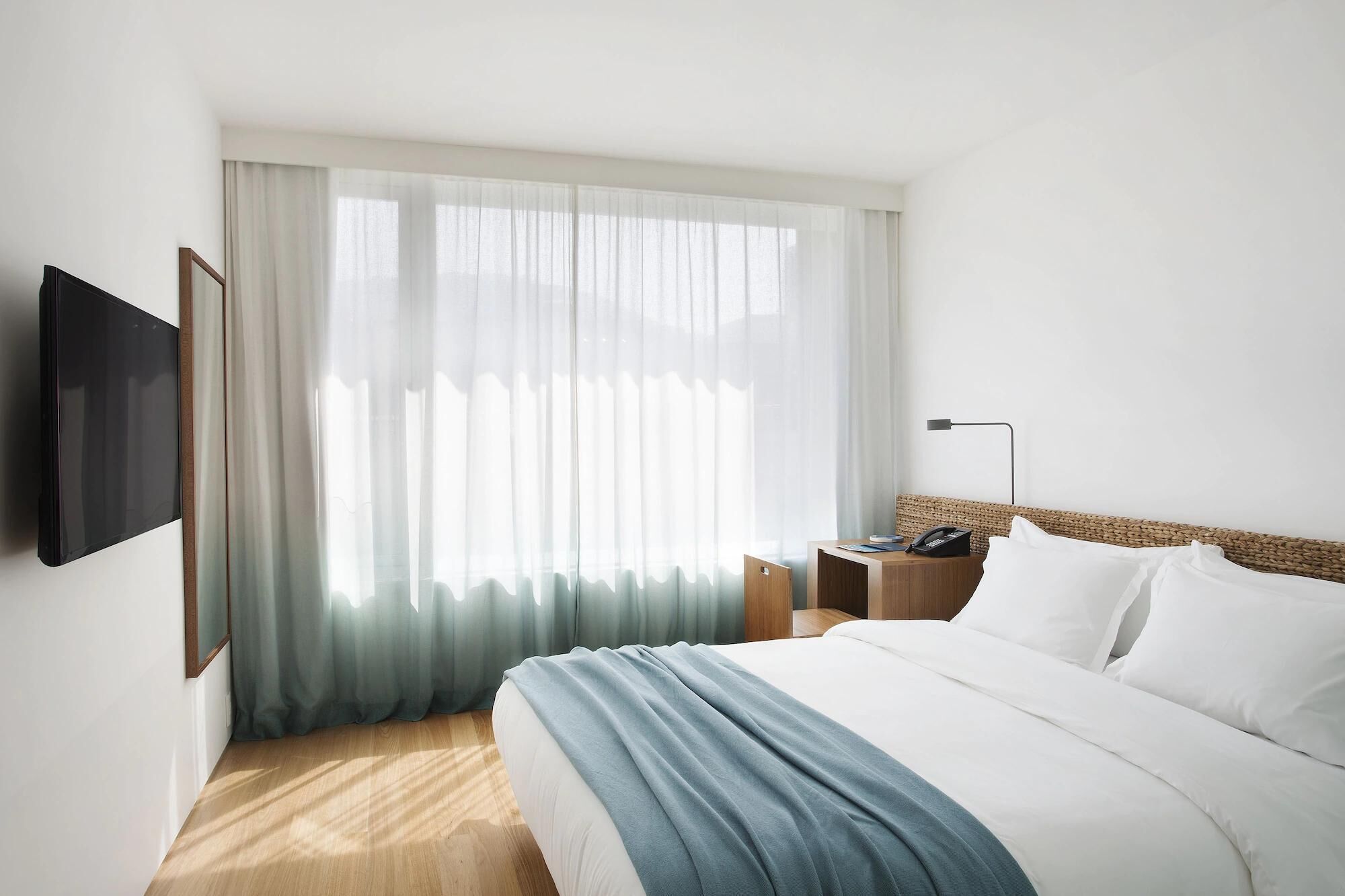 Une chambre d’hôtel avec un lit, une table de chevet avec une lampe, un téléviseur fixé au mur et des rideaux légers.