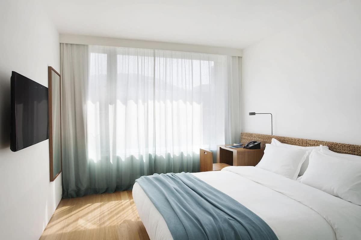 A hotel room with a bed, a bedside table with a lamp, a TV mounted on the wall, and sheer curtains.