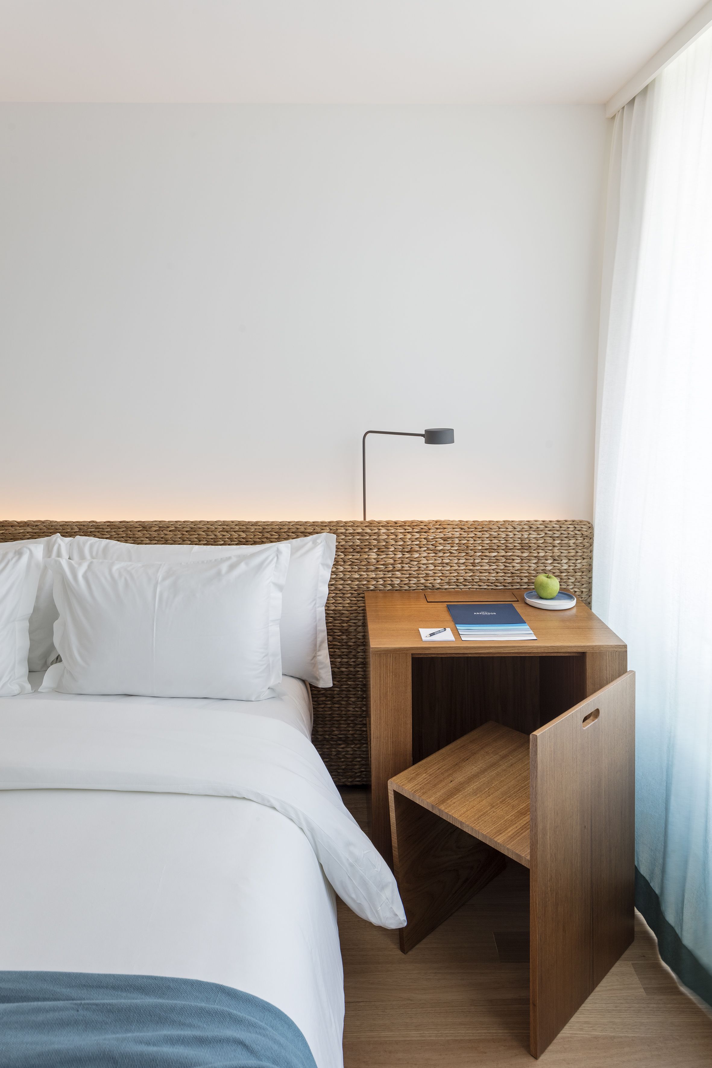 A neatly made bed with white bedding, a small wooden nightstand with a lamp, a book, and a fruit bowl.