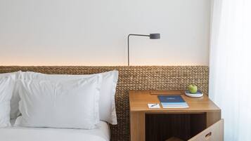 Een net opgemaakt bed met wit beddengoed, een klein houten nachtkastje met een lamp, een boek en een schaal fruit.