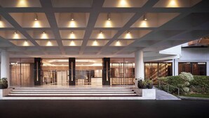 Une entrée d’hôtel moderne, avec un plafond au design géométrique, un escalier central et des portes vitrées donnant sur le hall d’accueil.