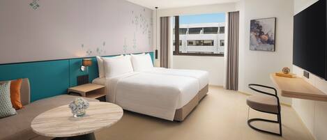 Habitación de hotel moderna con cama, sofá, una mesita con un florero, televisor y ventana con vistas a edificios.