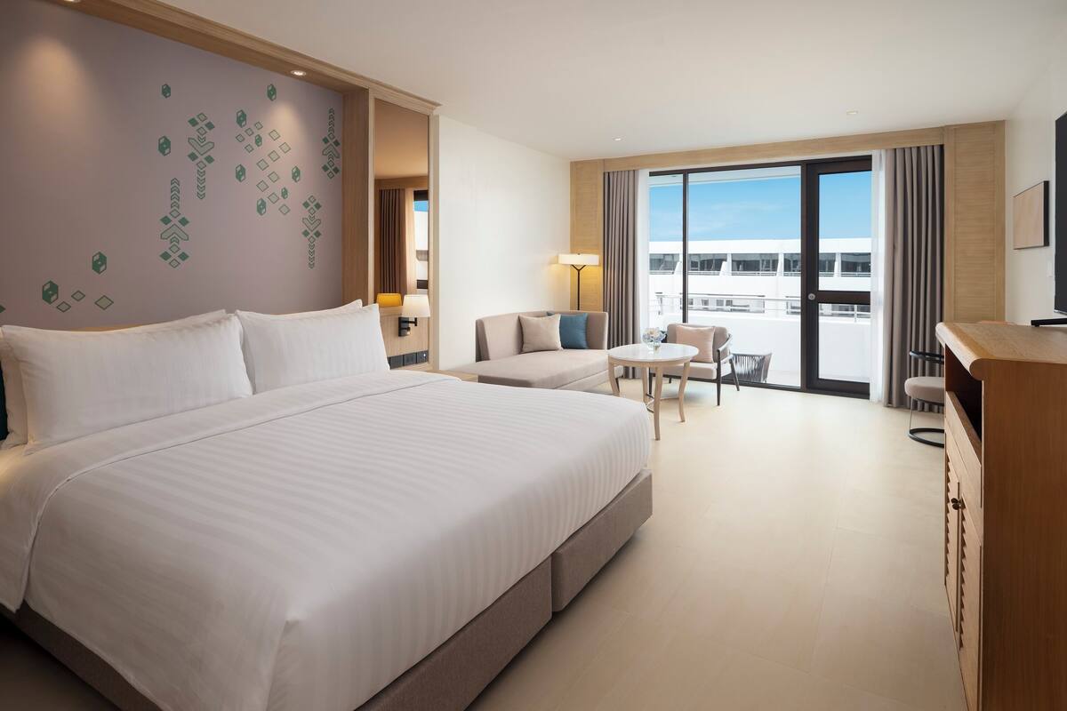 Habitación de hotel con una cama grande, un sofá, una mesita, una silla y un balcón con vistas.