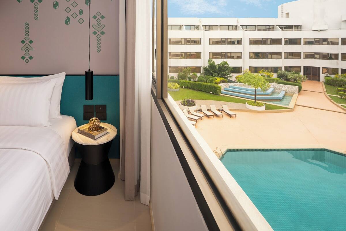 Habitación de hotel con cama, una mesita de noche con un libro, una ventana que da a una piscina y un edificio.