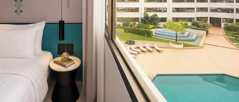 A hotel room with a bed, a small side table with a book, a window with a view of a swimming pool and a building.