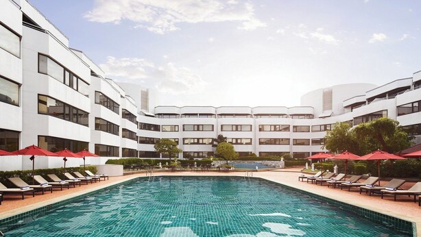 Un hôtel moderne doté d’une piscine centrale, d’espaces de détente agrémentés de parasols rouges et d’une façade au design symétrique.
