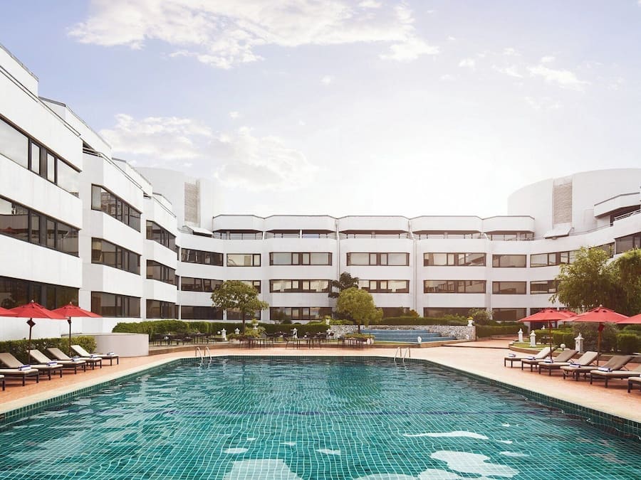 Un hotel moderno con una piscina central, zonas de descanso con sombrillas rojas y una fachada de edificio simétrica.