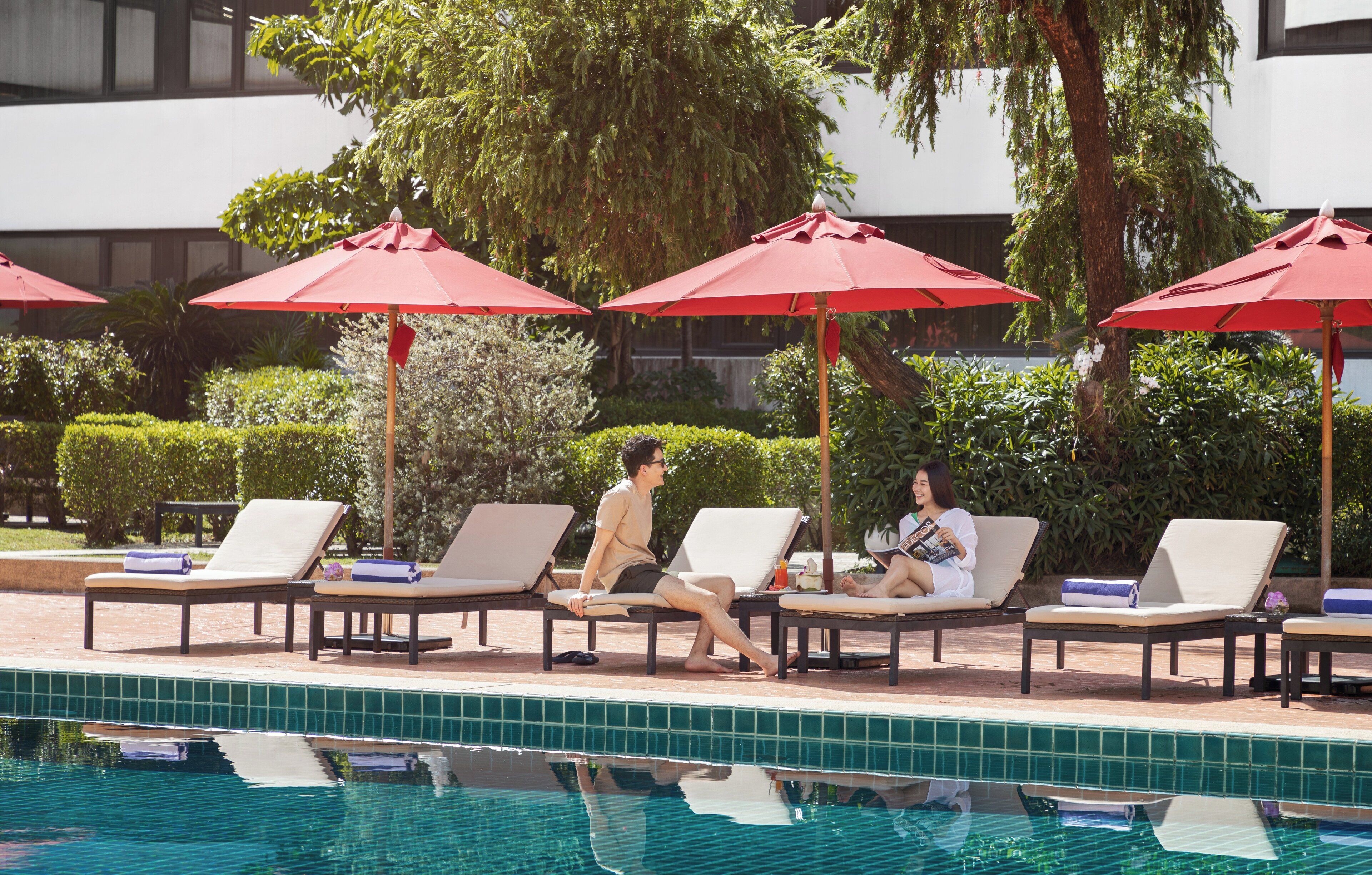 A poolside area with lounge chairs and red umbrellas, a tiled pool, and a building in the background.