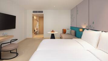 A hotel room with a bed, a TV, a desk with a chair, a small table with a lamp, and a wardrobe.
