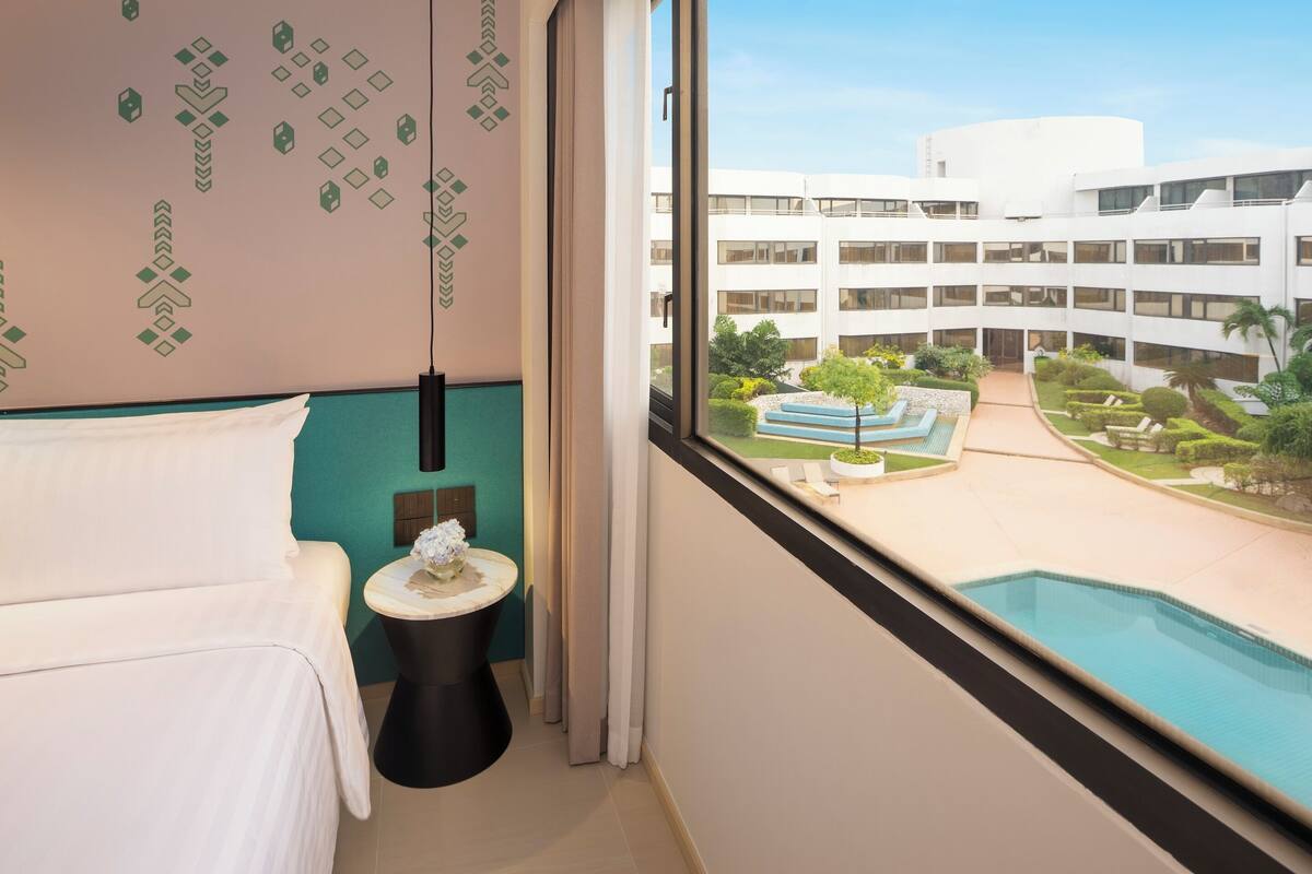 Habitación de hotel con cama, una mesita de noche con un florero, y ventana con vista a una piscina y un edificio.